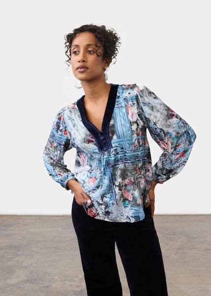 A woman modeling a long-sleeve button-up blouse with a blue, gray, and pink abstract painterly print, featuring a dark navy V-neck trim, paired with black pants against a plain studio background.