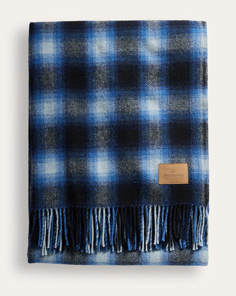 A folded plaid throw blanket in shades of blue, navy, and white, featuring a fringed edge and a small tan leather brand patch that reads 'The Pendleton'.