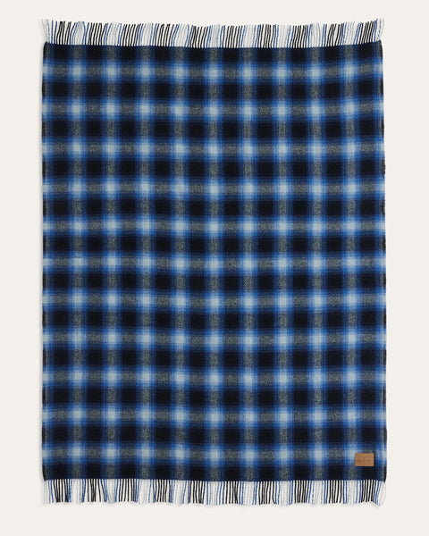 A rectangular plaid scarf with fringed edges, featuring a pattern of navy, light blue, and white. A small tan leather brand tag is visible in the bottom right corner, set against a solid off-white background.