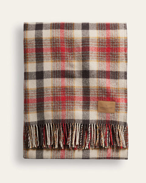 A folded plaid scarf with fringe, featuring a pattern of red, brown, black, and beige stripes. A small tan leather brand patch is visible on the right side, and the scarf is photographed against a light off-white background.