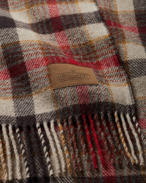 Close-up of a plaid wool blanket with a woven leather patch reading 'BENDLETON HOME PRODUCE' and fringed edges, showing cream, red, brown, and gray tones.