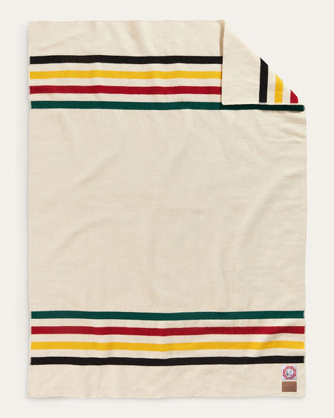 An off-white woven blanket with horizontal stripes in black, yellow, red, and green, shown flat with one corner folded over to reveal the reverse side and a small brand tag.