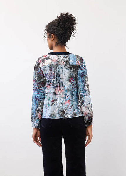 A woman viewed from behind wearing a long-sleeve blouse with a blue, gray, and pink abstract floral print, paired with black pants, against a plain white background.
