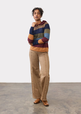 A model stands in a studio wearing a multicolor striped knit sweater with a hood, beige wide-leg pants, and brown shoes against a white wall and concrete floor.