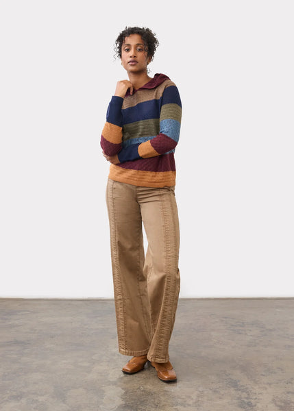 A model stands in a studio wearing a multicolor striped knit sweater with a hood, beige wide-leg pants, and brown shoes against a white wall and concrete floor.
