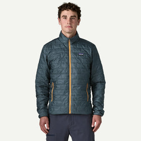 A man wearing a Patagonia quilted jacket in a slate blue-gray color with a mustard yellow zipper, standing against a light off-white background.