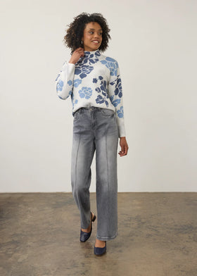 A woman modeling a white mock-neck knit sweater with a blue floral pattern, paired with wide-leg gray jeans and navy flats, standing against a plain off-white wall and concrete floor.