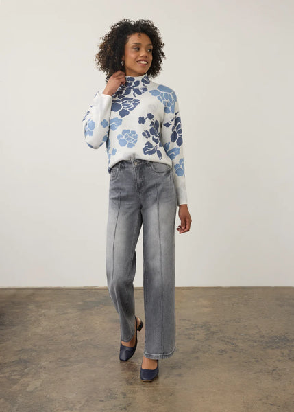 A woman modeling a white mock-neck knit sweater with a blue floral pattern, paired with wide-leg gray jeans and navy flats, standing against a plain off-white wall and concrete floor.