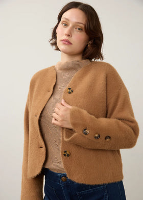 A woman wearing a cropped, fuzzy camel-brown cardigan over a beige ribbed turtleneck sweater and dark blue jeans, photographed against a plain light gray studio background.
