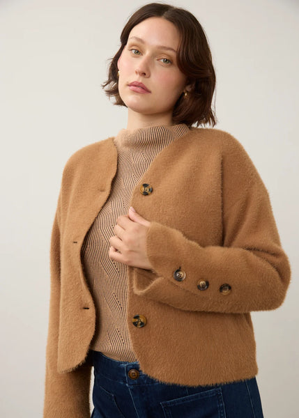 A woman wearing a cropped, fuzzy camel-brown cardigan over a beige ribbed turtleneck sweater and dark blue jeans, photographed against a plain light gray studio background.