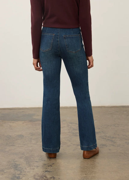 A person shown from the back wearing blue flare jeans, a burgundy long-sleeve top, and brown shoes in a studio setting with a plain light wall and concrete floor.