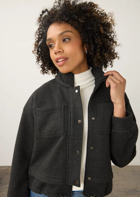 A woman with curly dark hair wearing a dark charcoal gray button-up jacket over a white ribbed turtleneck sweater and blue jeans, photographed against a plain light gray studio background.