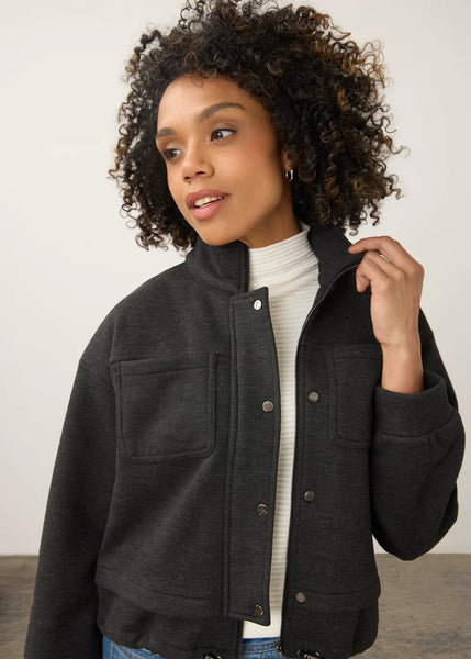 A woman with curly dark hair wearing a dark charcoal gray button-up jacket over a white ribbed turtleneck sweater and blue jeans, photographed against a plain light gray studio background.
