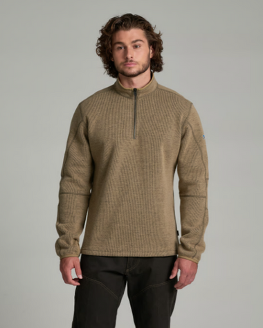 A man wearing a textured quarter-zip pullover sweater in a warm khaki-tan color, paired with black pants, standing against a solid light gray studio background.