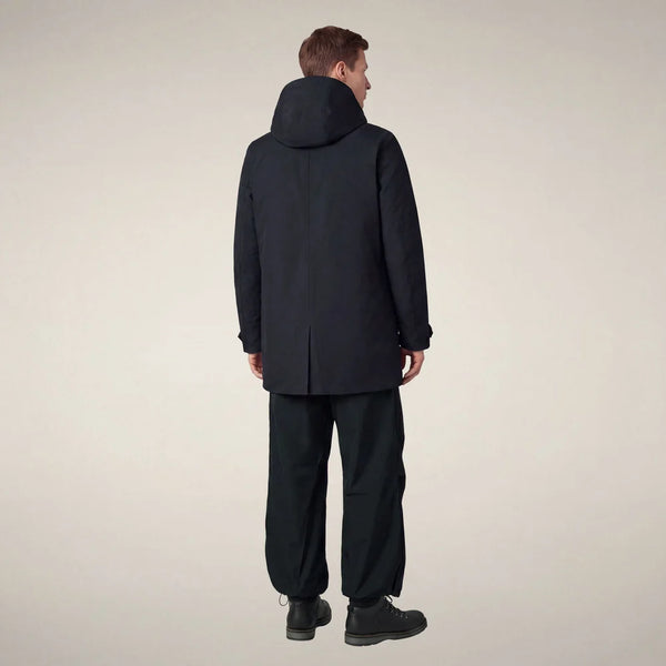 A man viewed from the back wearing a dark navy hooded jacket and matching dark pants against a plain light beige studio background.