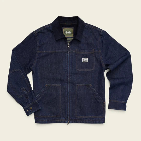 A dark indigo denim work jacket with a zippered front, chest pocket, and elbow patches, laid flat on a light beige background.