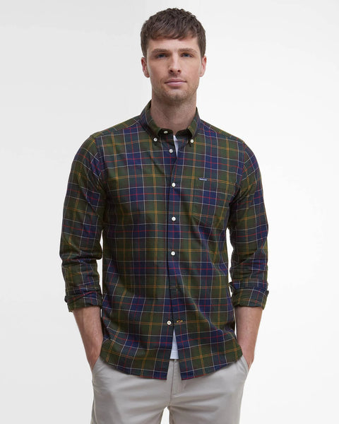 A man wearing a long-sleeve button-down plaid shirt in green, blue, and yellow, paired with light beige trousers, standing against a plain white studio background.