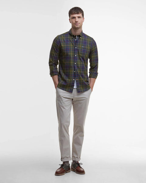 A man modeling a long-sleeve button-down plaid shirt in green, purple, and yellow, paired with beige trousers and brown shoes, against a plain white studio background.