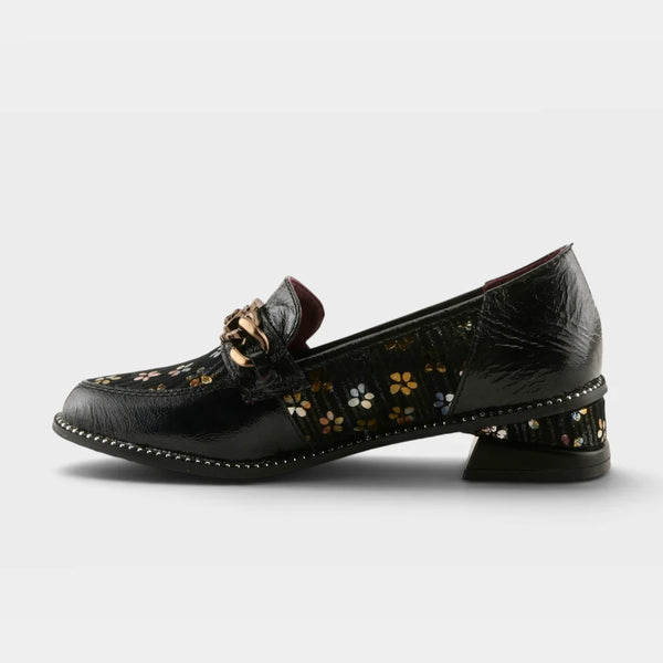 A single black leather loafer with a low block heel, featuring a floral pattern on the sides and a gold-tone metal buckle detail on the vamp, shown against a light gray background.