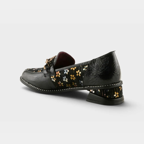 A black leather loafer with a low block heel, featuring a floral patterned insole and a decorative gold-tone bow on the vamp, photographed from a side angle against a light gray background.