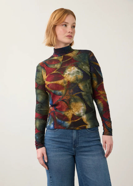 A woman wearing a long-sleeve mock neck top with an abstract multicolor print in red, green, yellow, and blue, paired with blue jeans, photographed against a plain light gray background.
