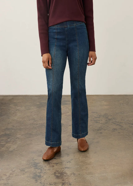 A person wearing blue straight-leg denim jeans, a burgundy long-sleeve top, and brown leather shoes, standing against a plain off-white wall and concrete floor.