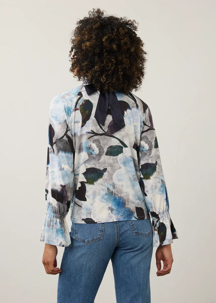 A woman viewed from behind wearing a long-sleeve blouse with a blue, black, and white abstract floral print and a dark bow at the back of the neck, paired with blue jeans against a plain light background.