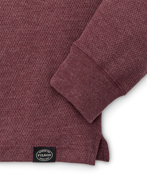 Close-up of a ribbed knit crewneck sweatshirt in a heathered dusty rose or mauve color, showing the ribbed cuff and a black sewn-on brand label that reads 'PILSON Since 1967'.