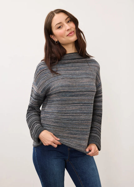 A woman wearing a gray and blue heathered striped long-sleeve mock neck sweater with blue jeans, standing against a plain off-white background.