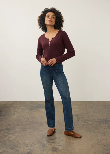 A woman modeling a long-sleeve burgundy henley top with a split neckline and silver zipper detail, paired with blue jeans and brown leather shoes, standing against a plain off-white wall.