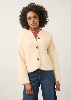 A woman with curly dark hair wearing a cream-colored fuzzy cropped cardigan with brown buttons, a red and white patterned turtleneck, and blue jeans, standing against a plain off-white studio background.