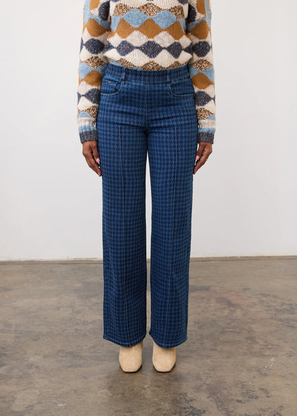 A person wearing blue houndstooth wide-leg trousers, a multicolor argyle sweater, and beige pointed-toe heels, standing against a plain white wall and concrete floor.