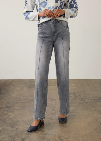 A model wearing light gray washed denim jeans with front seam detailing, paired with a blue and white patterned sweater and navy blue heels, standing against a neutral studio backdrop.