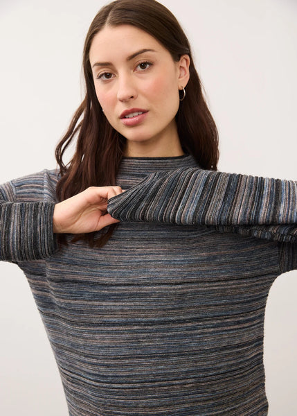 A woman wearing a ribbed knit mock neck sweater with horizontal stripes in blue, gray, and brown tones, holding up the sleeve to show the texture.
