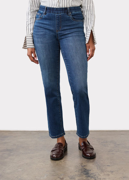 A person wearing blue straight-leg denim jeans, a black and white striped long-sleeve top, and dark brown leather loafers, standing against a white studio wall.