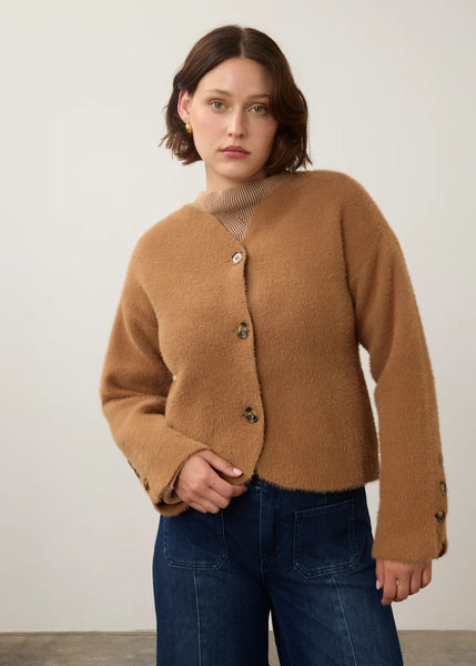 A woman wearing a cropped, fuzzy camel-brown cardigan with dark buttons over a beige ribbed turtleneck, paired with dark blue jeans, standing against a plain off-white wall.