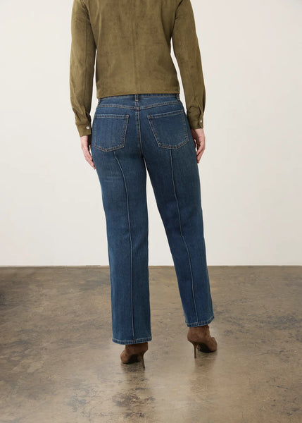 A person shown from the back wearing dark blue straight-leg jeans, an olive green long-sleeve top, and brown heeled boots against a plain off-white wall and concrete floor.