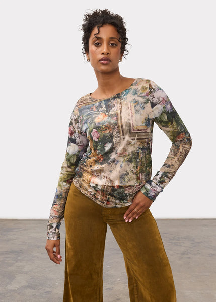 A woman modeling a long-sleeve top with a vintage, multicolored scenic print, paired with wide-leg mustard brown trousers, against a neutral studio background.