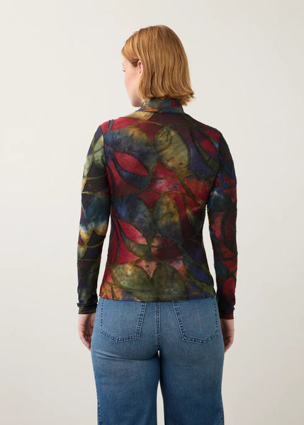 A woman viewed from the back wearing a long-sleeve mock-neck top with an abstract multicolor print in red, blue, green, and yellow, paired with blue jeans, against a plain light gray background.