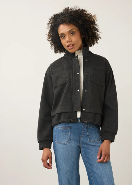 A woman with curly dark hair wearing a dark charcoal gray cropped button-up jacket over a white ribbed turtleneck and blue jeans, standing against a plain light gray studio background.