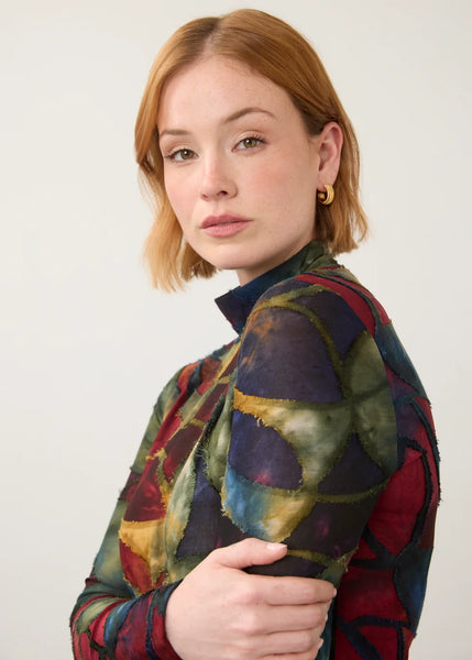 A woman with short auburn hair wearing a colorful patchwork-patterned long-sleeve top and small gold hoop earrings, photographed against a plain light gray studio background.