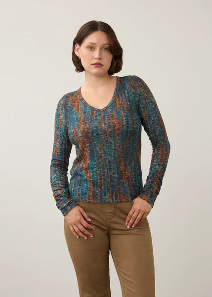 A woman modeling a long-sleeve V-neck knit top with a vertical textured pattern in teal blue, rust orange, and brown, paired with tan pants, against a plain light gray studio background.