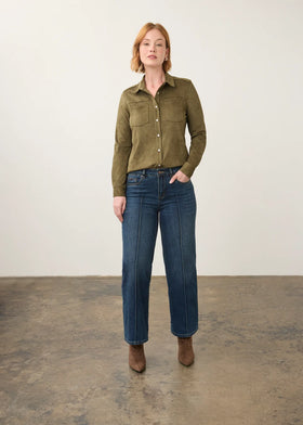 A woman stands in a studio wearing an olive green long-sleeve button-up shirt, dark blue straight-leg jeans, and brown ankle boots against a plain off-white wall and concrete floor.