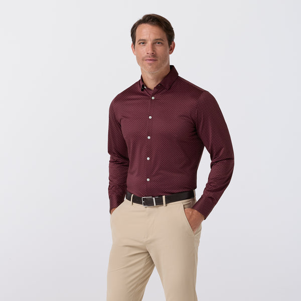 A man wearing a burgundy long-sleeve button-down dress shirt with a subtle micro-dot pattern, paired with beige trousers and a black belt, standing against a light gray studio background.