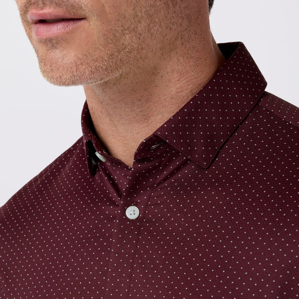 Close-up of a man wearing a burgundy polo shirt with a small white micro-dot pattern, showing the collar, button, and part of his lower face against a light gray background.