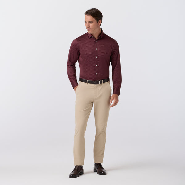 A man modeling a long-sleeve burgundy button-down dress shirt with beige dress pants, a black belt, and dark brown dress shoes, photographed against a light gray studio background.