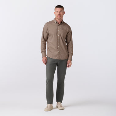 A man modeling a brown long-sleeve button-down shirt with dark gray pants and beige sneakers, standing against a light gray studio background.