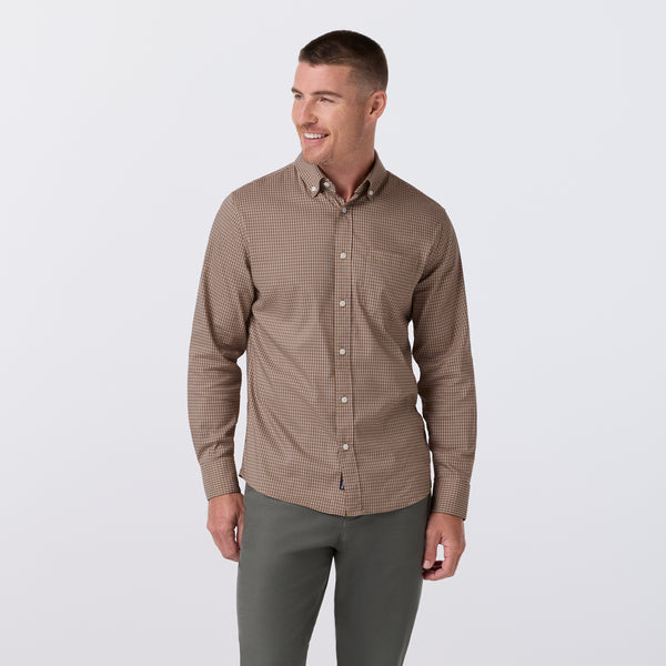 A man wearing a brown and beige micro-check patterned long-sleeve button-down shirt with dark gray pants, standing against a light gray studio background.