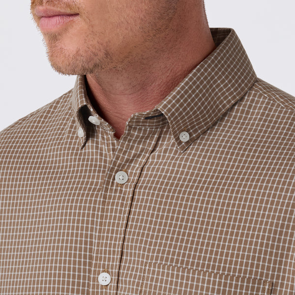 Close-up of a man wearing a brown and beige checkered button-down shirt, showing the collar, buttons, and chest area against a light gray background.