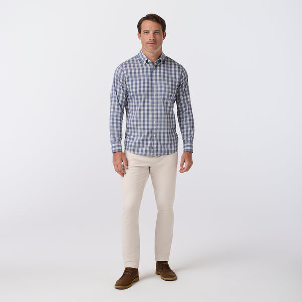A man modeling a blue and white checkered long-sleeve button-down shirt, paired with beige pants and brown shoes, standing against a light gray studio background.